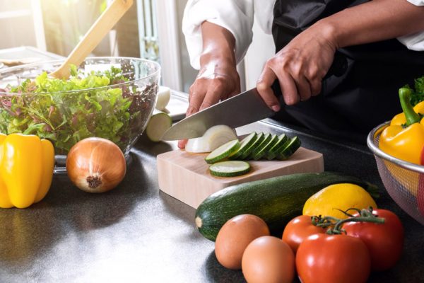 Close up of woman hand cutting onion on chopping wood board with sharp knife and cooking vegetables salad in kitchen.